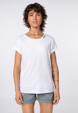 Woman with curly hair wearing a white short-sleeve shirt with buttons near the collar and green rolled-up shorts, standing against white background.