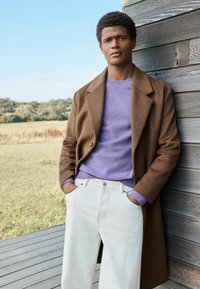Brauner Wollmantel über einem lila Strickpullover, kombiniert mit weißen Jeans. Der Mantel hat einen Reverskragen und einen einzelnen Knopfverschluss.