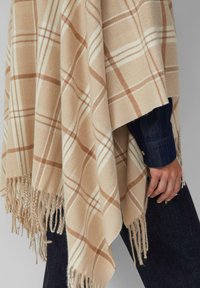 Beżowy pledowy poncho w brązowy wzór w kratę, z frędzlami na brzegach, wykonane z miękkiego materiału, noszone na ciemnoniebieskim rękawie dżinsowym.
