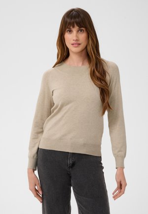Jeune femme aux longs cheveux bruns portant un pull beige et un jean gris foncé, debout devant un fond gris clair.