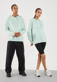 Piparmētru zaļas kapuces ar kangaru kabatām, savienotas ar melniem plašiem biksēm un melnām legingām. Strukturēti sporta apavi. Vienkāršs, ikdienišķs dizains.