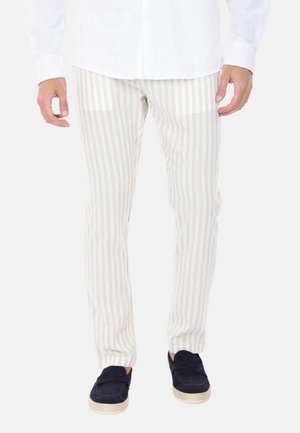 Uomo che indossa pantaloni beige e bianchi a righe verticali, camicia bianca e scarpe slip-on blu scuro, in piedi su uno sfondo semplice.