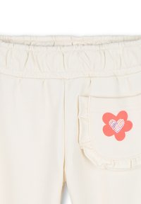 Crème-kleurige broek met een elastische tailleband. Voorzien van een decoratieve zak met geplooid randje en een roze bloemenafbeelding met een hartontwerp.