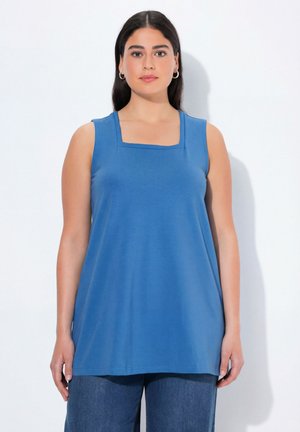 Kobieta z długimi ciemnymi włosami, ubrana w bezrękawnik w niebieskim kolorze z kwadratowym dekoltem i niebieskie dżinsy, stojąca na białym tle.