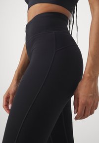 Leggings noirs taille haute en tissu extensible, présentant une texture lisse, des coutures latérales et une coupe près du corps.
