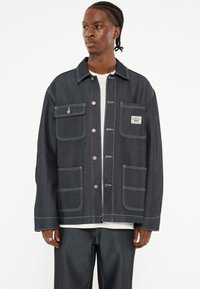 Donkerdenimjas met een knoopsluiting aan de voorkant, twee borstzakken en twee zakken onderaan. Witte stiksels als accent en een label op de borstzak.