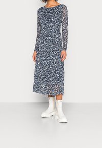 Sukienka z długimi rękawami w kolorze granatowym, w białe plamki, z talią podkreśloną w pasie i zwiewnym dołem. Połączona z masywnymi białymi botkami za kostkę.