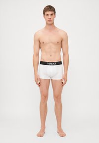 Fehér pamut boxer alsó fekete elasztikus derékkal, amelyen a "Versace" logó található. Sima textúra, testhez álló kialakítás és combközép hosszúság.