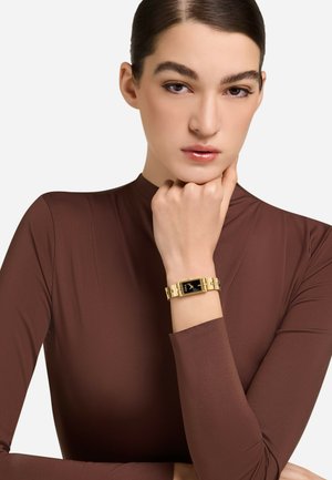 Zlata ura z pravokotnim rezom, črnim številčnico in zlatim povezovalnim trakom. Model nosi prilegajočo rjavo majico in se opira s brado na roko.