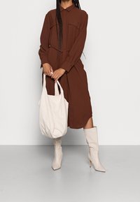 Robe longue marron à manches longues avec deux poches poitrine, taille cintrée et une coupe fluide. Associée à des bottes beiges jusqu'aux genoux et un sac fourre-tout crème.