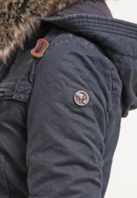 Marinblå parkajacka med en fuskpäls-hood, sidfickor och en rund logotypapplikation på vänster arm. Strukturerad, hållbar tyg.