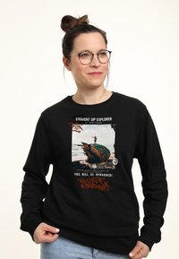 Felpa nera con un design grafico che presenta una scena di spiaggia, elementi testuali e accenti in stile graffiti arancioni e bianchi.