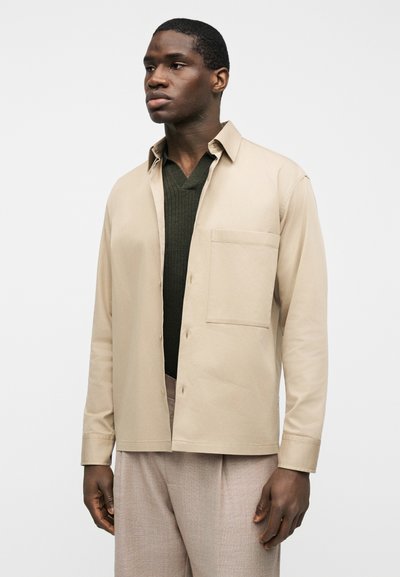 Νέος άνδρας που φοράει μπεζ πουκάμισο με κουμπιά πάνω από σκούφο πράσινο πουλόβερ με V λαιμόκοψη και ανοιχτό καφέ παντελόνια, στέκεται μπροστά σε λευκό φόντο.