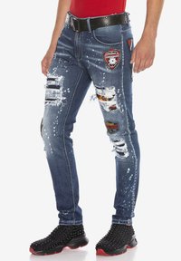 Jean en denim bleu foncé avec des déchirures usées et des éclaboussures de peinture blanche. Comprend despatches et une ceinture noire. Design ajusté avec un léger stretch.