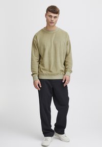 Olivengrønn hettegenser med løs passform, ribbede mansjetter og hem, kombinert med mørke bukser. Modellen har på seg hvite sneakers. Ensfarget bakgrunn.