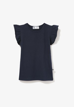 Uśmiechnięta młoda dziewczyna ubrana w granatowy top z falbankami na rękawach, białą plisowaną spódnicę i srebrne buty typu Mary Jane, stoi na jednolitym tle.