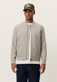 Γκρι bomber μπουφάν με φερμουάρ μπροστά, πλαϊνές τσέπες και μανσέτες με ριμπ ύφανση. Φορώντας από πάνω λευκό πουκάμισο, συνδυασμένο με σκουρόχρωμο τζιν και ελιά καπέλο.