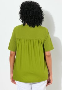 Top verde a maniche corte realizzato in tessuto leggero, con un design a pieghe sulla schiena, scollatura tonda e vestibilità rilassata.