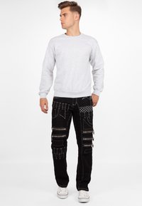 Felpa grigia a maniche lunghe abbinata a pantaloni cargo neri con più zip e dettagli testurizzati. Sneakers bianche completano il look.