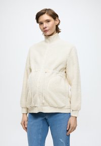 Giacca in pile color crema con collo alto, vita con coulisse e tasche laterali; presenta una superficie testurizzata e polsini a coste.