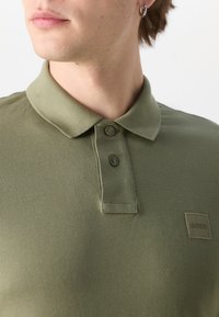 Jeune personne portant un polo vert olive avec deux boutons et un petit logo carré BOSS sur la poitrine, légèrement orientée vers la gauche.