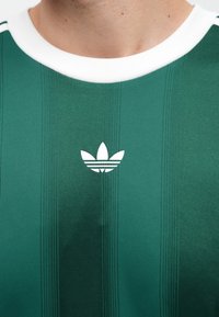 Muškarac u zelenoj Adidas sportskoj majici s bijelim ovratnikom i Adidas logom na sredini prsa.