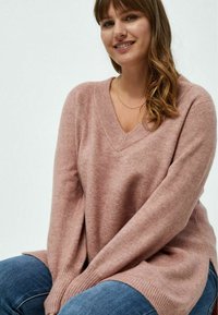 Rosa V-Ausschnitt-Pullover aus weichem Material; mit langen Ärmeln, gerippten Bündchen und seitlichen Schlitzen; kombiniert mit Jeans.