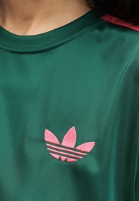 Πράσινο αθλητικό πουκάμισο με ροζ κεντημένο λογότυπο τριφυλλιού Adidas στο στήθος, φορεμένο από άτομο με μακριά σκούρα μαλλιά.