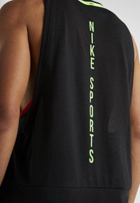 Lähikuva henkilöstä, joka käyttää mustaa hihattomaa urheilupaitaa, jossa on pystysuunnassa limevihreää "NIKE SPORTS" -tekstiä selässä.