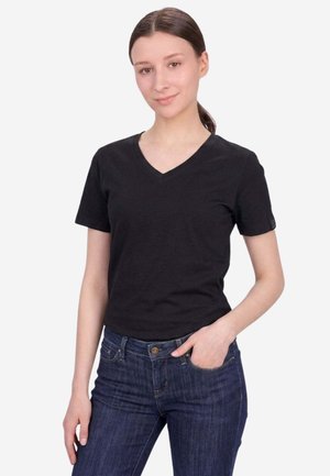 Giovane donna con capelli castani legati dietro, che indossa una maglietta nera con scollo a V e maniche corte e jeans blu, una mano in tasca, sorride dolcemente.