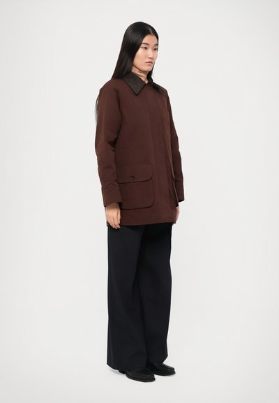 Cască maro cu guler, două buzunare frontale și un nasture. Asortată cu pantaloni largi negri și pantofi negri. Textură netedă, design modern.