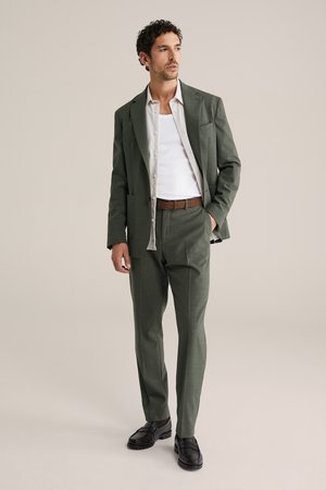 Man klädd i en grön kostym med en vit öppen skjorta över en vit linne, brunt skärp och svarta loafers, stående med ena handen i fickan.