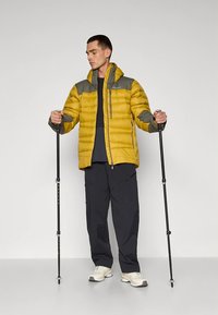 Yellow puffer jacket with gray accents, zippered front, adjustable hood, and two side pockets. Model holds trekking poles, wearing dark pants.
