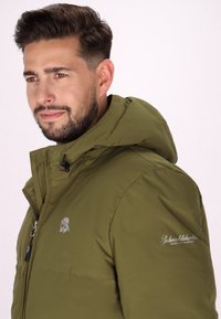 Giacca isolante verde oliva con cappuccio, caratterizzata da una texture liscia, chiusura con zip e un logo sul petto e sulla manica.