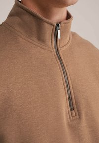 Pullover marrone con zip a un quarto, realizzato in tessuto morbido. Caratterizzato da un colletto alto e una zip metallica con un tirante rettangolare. Texture liscia.