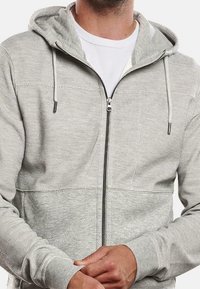 Sweat à capuche gris à fermeture éclair, fabriqué en tissu texturé avec une partie inférieure unie, cordons au niveau de la capuche et fermeture éclair lisse.