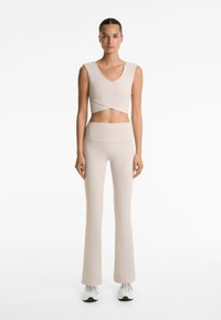 Beige idrotts crop top med V-ringning och korsettdesign, parat med högmidjade beige utsvängda leggings. Slät textur, minimal detaljer.
