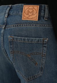 Blå denimjeans med en lærrem på livet med en preget logo. Kontraststing på baklommene i et buet design.