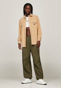 Camisa de botones color marrón con bolsillo en el pecho, pantalones relajados color verde oliva y zapatillas de piel blancas. El modelo está de pie mirando hacia adelante sobre un fondo neutro.