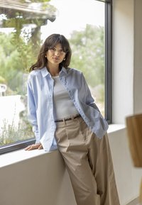 Femme appuyée contre un rebord de fenêtre, portant une chemise bleu clair à boutons, un haut blanc, un pantalon beige, avec de la verdure visible à l'extérieur de la fenêtre.