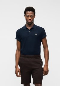 Homme portant un polo bleu marine à manches courtes avec un petit logo vert et un short marron foncé jusqu'aux genoux, debout devant un fond blanc.
