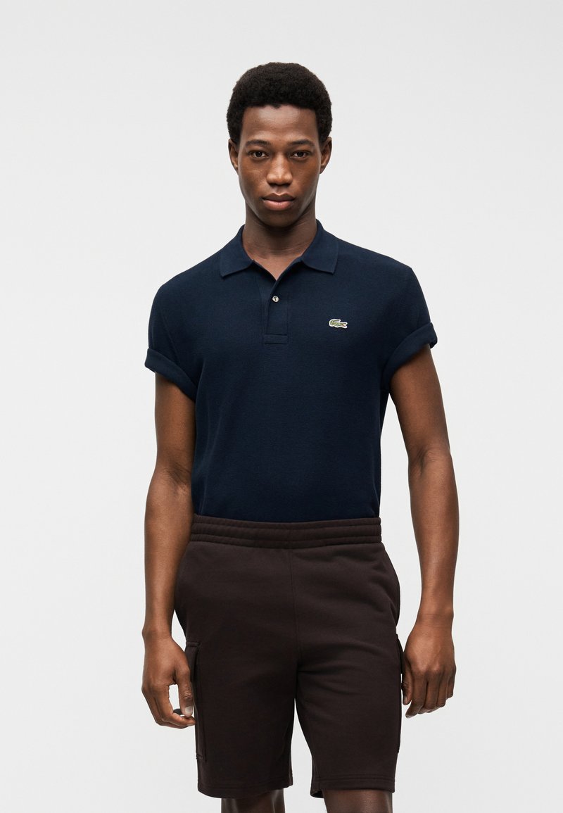 Homme portant un polo bleu marine à manches courtes avec un petit logo vert et un short marron foncé jusqu'aux genoux, debout devant un fond blanc.
