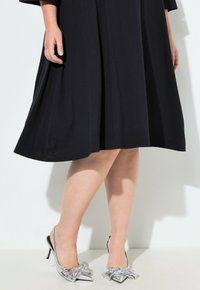 Eine Nahaufnahme des Unterkörpers einer Frau in einem schwarzen Kleid, die glänzende silberne High Heels mit Schleifen trägt, vor einem hellen Hintergrund.
