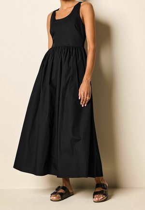 Femme portant une robe maxi noire sans manches avec une taille froncée, associée à des sandales noires à double boucle, debout contre un mur beige.