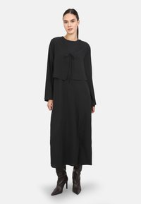 Schwarzes langes Kleid mit einer kurz geschnittenen, gebundenen Overlay. Besondere Merkmale sind lange Ärmel, eine lockere Passform und eine weiche, glatte Textur. Kombiniert mit Stiefeln mit Absatz.