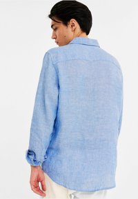 Himmelblaues Leinenhemd mit lockerer Passform, langen Ärmeln und knöpfbarem Bündchen. Weiche Textur und abgerundeter Saum hinten.