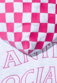 Różowo-biała materiałowa tkanina w kratkę o miękkiej teksturze, ułożona na szarym materiale z różowymi haftowanymi literami. Zauważalne kontrastujące wzory.