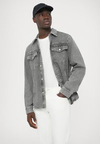 Γκρι τζιν μπουφάν με δύο τσέπες στο στήθος, κλείσιμο με κουμπιά και μακριά μανίκια, σε συνδυασμό με λευκό μπλουζάκι και λευκό παντελόνι. Φοράει μαύρο καπέλο.