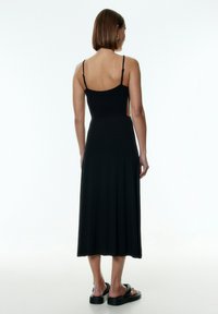 Schwarzes Midi-Kleid mit dünnen Trägern und Seitenausschnitten. Hergestellt aus weichem Stoff, verfügt es über ein eng anliegendes Oberteil und einen ausgestellten Rock. Dazu werden schwarze Sandalen getragen.