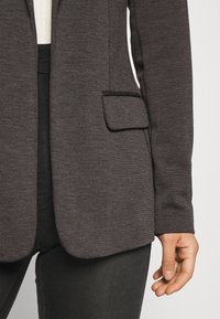 Donkergrijze, gestructureerde blazer met een enkele zak, gecombineerd met zwarte jeans, met nadruk op een gestroomlijnd ontwerp en contrasterende materialen.